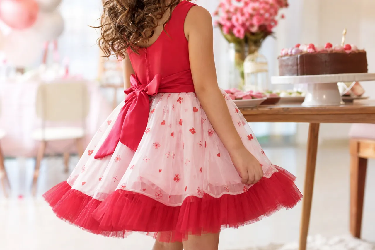 Menina sem rosto identificável usando vestido infantil vermelho e rosa em cenário de aniversário