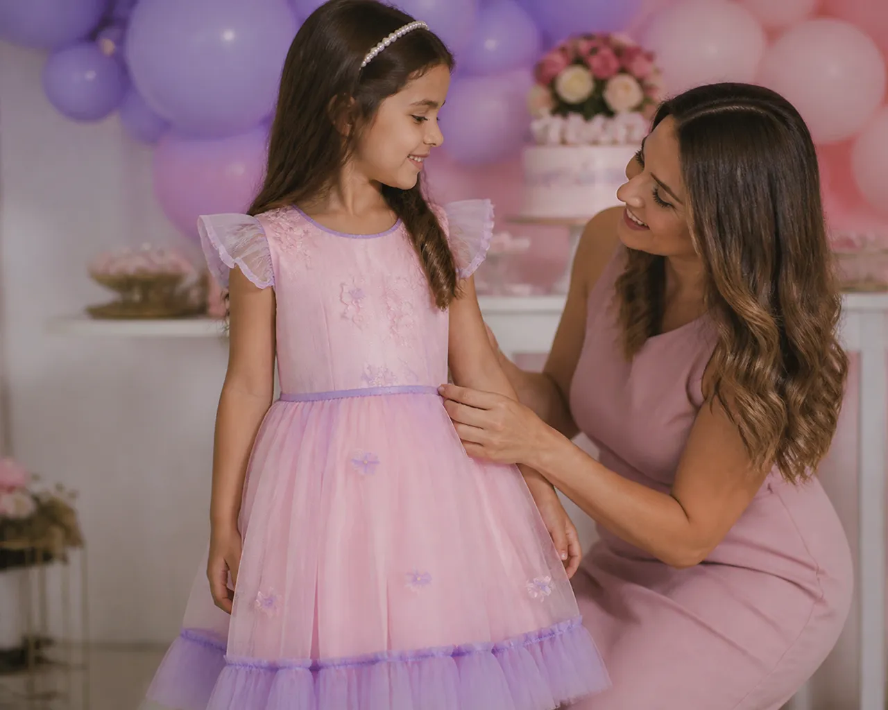 Menina usando vestido infantil rosa e lilás em festa de aniversário com balões enquanto a mãe confere o caimento em Nova Crixás
