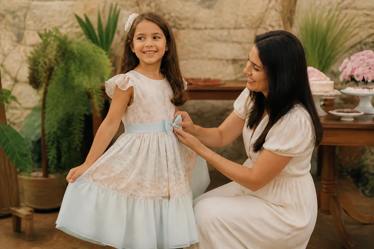 Menina usando vestido infantil de festa enquanto a mãe ajusta o laço para conferir caimento e conforto